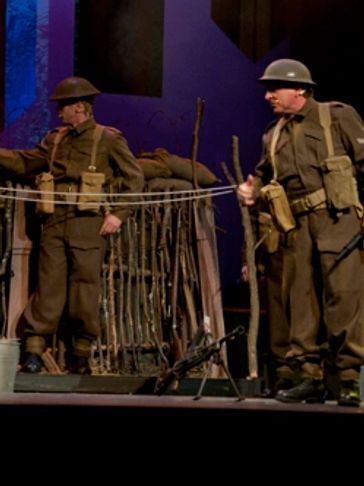 Two soldiers in vintage uniforms standing near a makeshift barricade on stage.