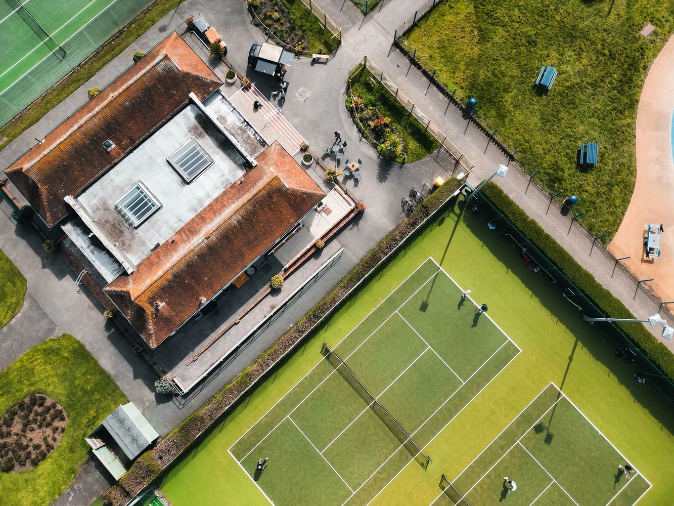 Aerial view of a tennis court beside a building with a red roof and surrounding greenery.