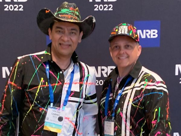 Two men in colorful splattered shirts and hats at NRB 2022 event.