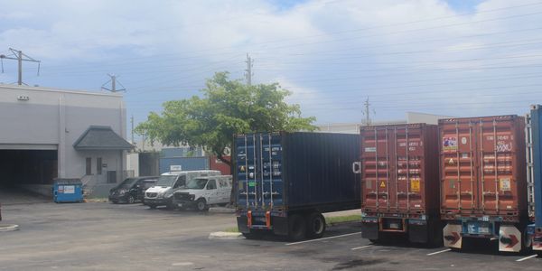 Shipping containers parked in an industrial lot next to a warehouse and vans.