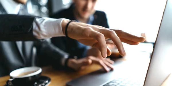 A person pointing at a laptop screen during a meeting with coffee nearby.