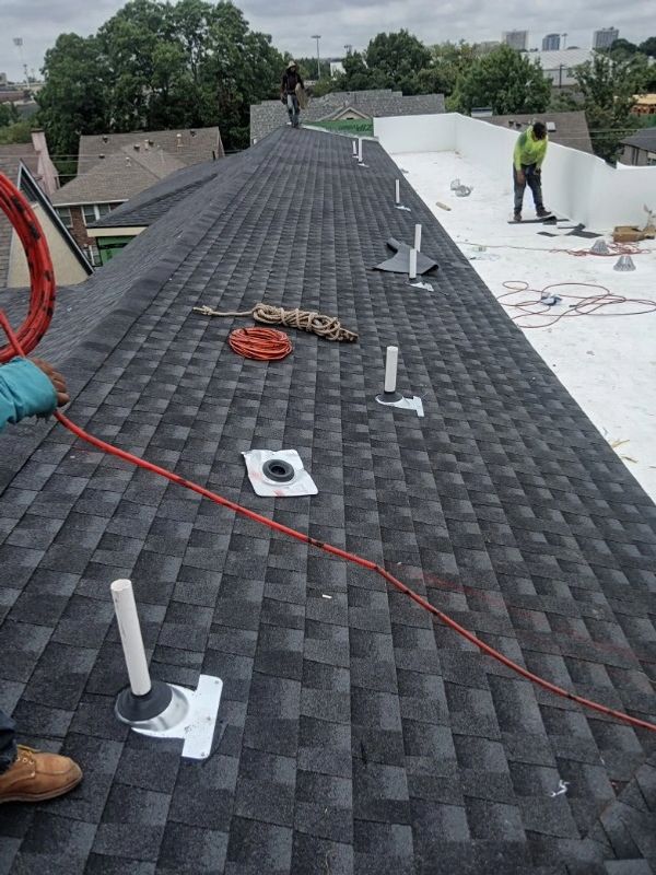 Workers installing black asphalt shingles on a roof with pipes and tools scattered.