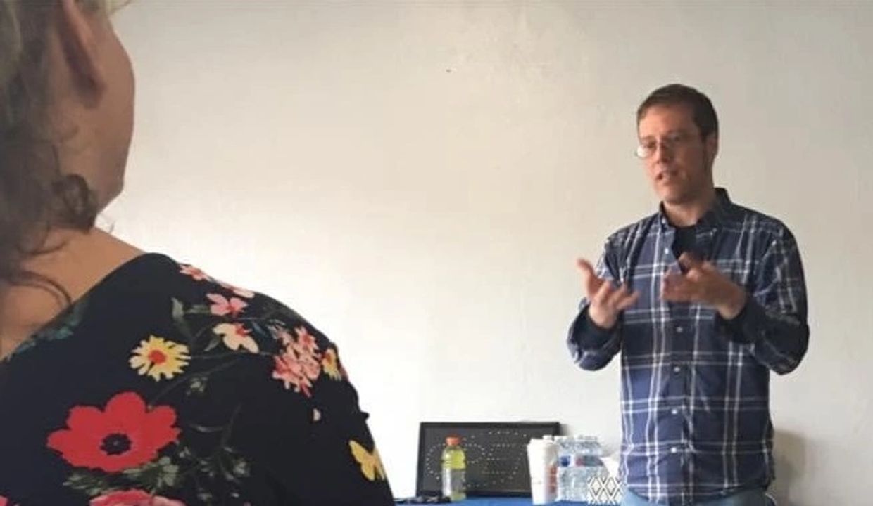 Man in plaid shirt speaking to a woman in floral dress in a casual setting.