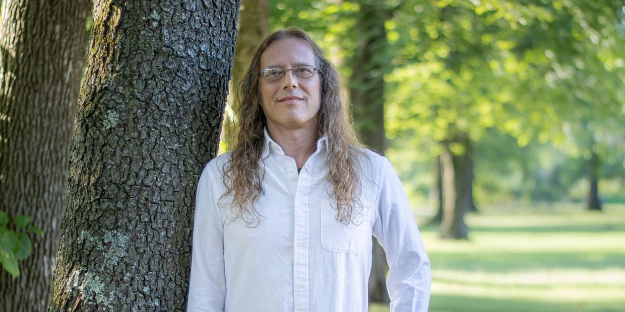 Man with long hair and glasses leaning against a tree in a sunlit park.