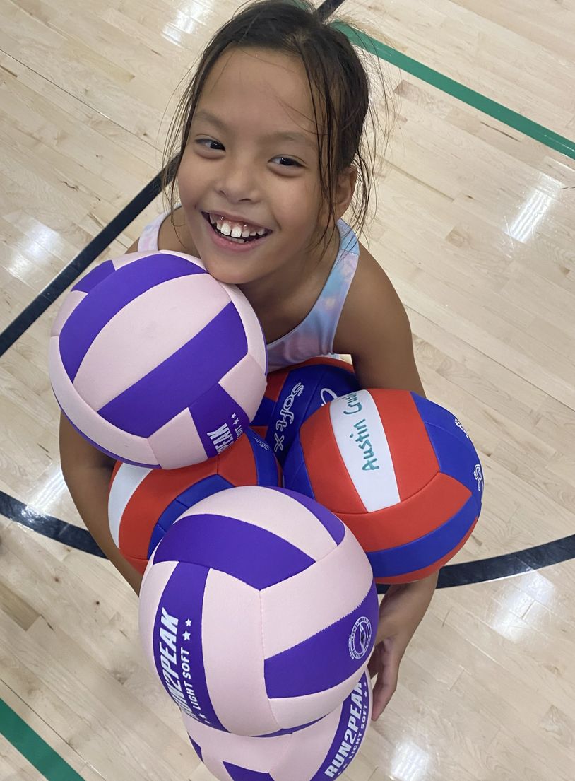Happy girl holding multiple colorful volleyballs on a gym floor.