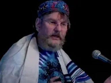 Man with beard speaking into microphone wearing a patterned cap and shawl.
