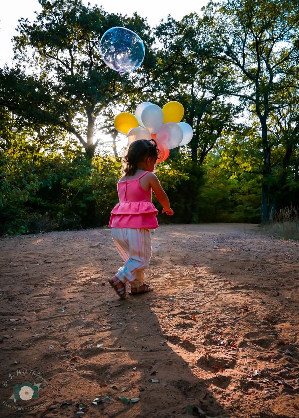 Nature scenery. Toddler wearing pink yellow and white outfit, holding matching balloons.