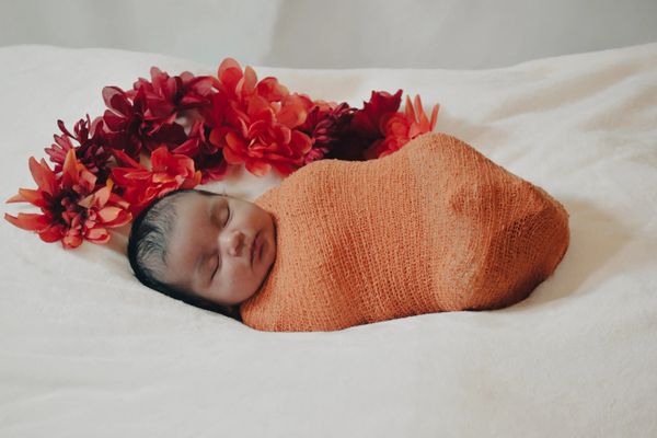 Infant in orange wrap, surrounded by red and orange flowers with soft white background