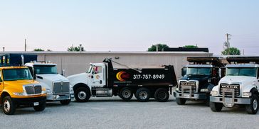 A lineup of five colorful dump trucks parked on gravel in front of a warehouse.