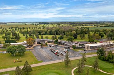 A large building with parking surrounded by lush green fields and trees under a blue sky.