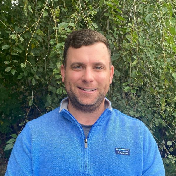 Smiling man in a blue jacket standing outdoors with greenery in the background.