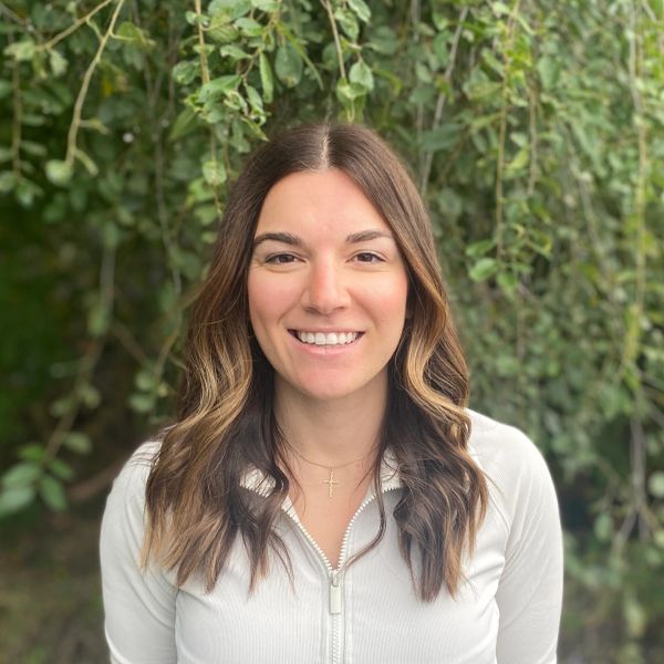 Smiling woman with wavy hair standing outdoors with greenery in the background.