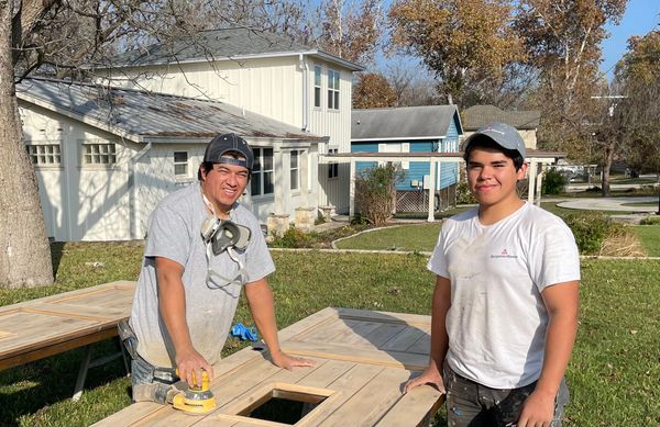 Two men working on wooden panels outdoors on a sunny day.