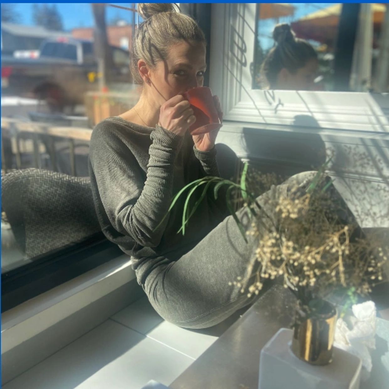 Woman in cozy grey outfit sipping from a red cup by a window seat.