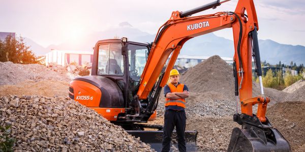 Construction worker standing next to an orange Kubota excavator on a gravel site.