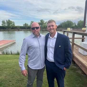 Two men posing by a lakeside, one in a suit and the other in a casual shirt.