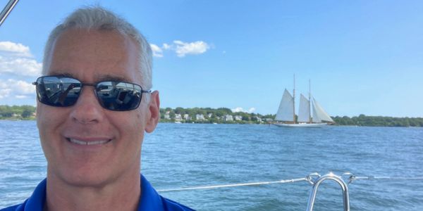 Man wearing sunglasses on a boat with a sailboat in the background.