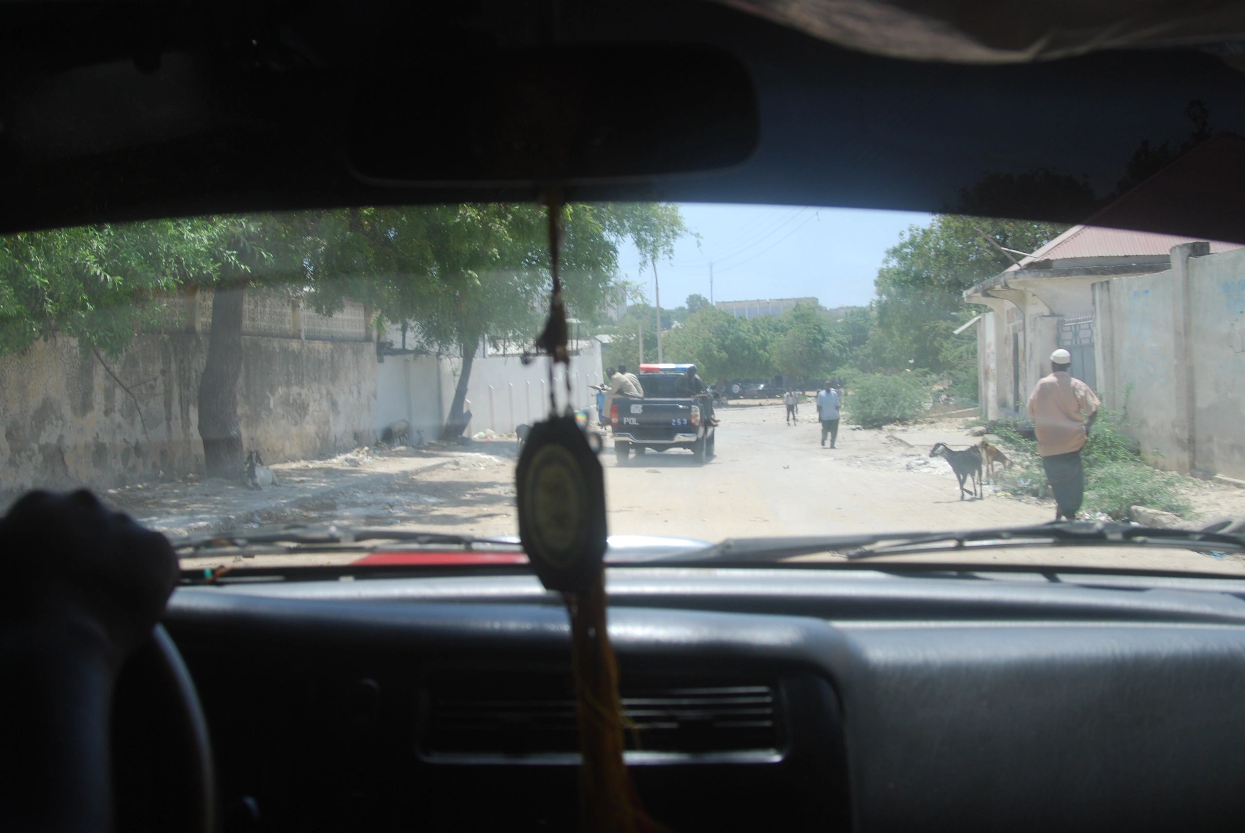 View through a car windshield of a police vehicle on a dirt road with pedestrians and goats.