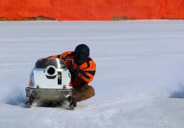 Person riding a snowmobile on a snowy surface wearing an orange jacket.
