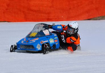 Racer in orange gear speeds on blue snowmobile on snowy track.