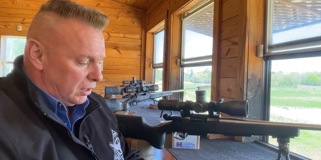 Man in a shooting range with rifles and scopes beside him.