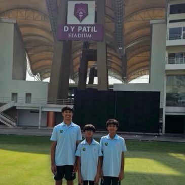 Three boys in blue shirts standing on green grass at DY Patil Stadium.
