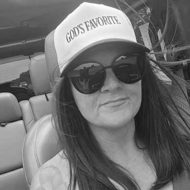 Woman wearing a hat that says 'GOD'S FAVORITE' and large sunglasses in a car.