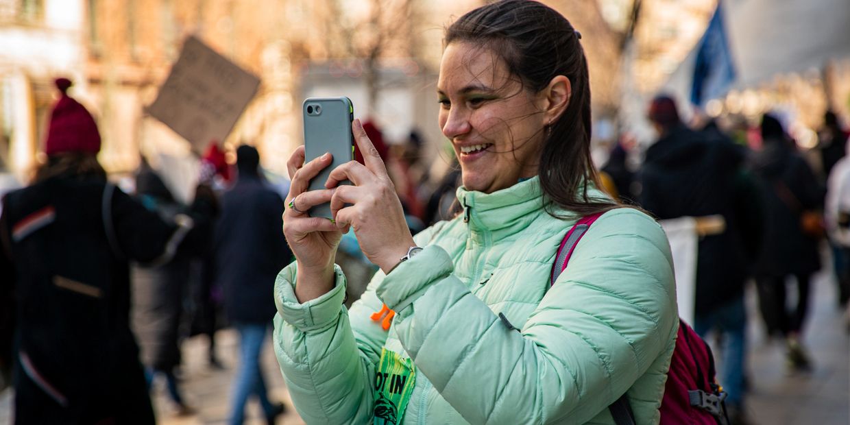 A woman in a green jacket smiling and taking a photo with her phone at an outdoor event.