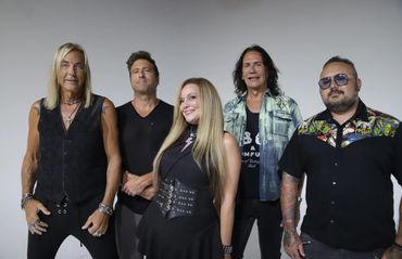 A diverse rock band posing together in front of a gray backdrop.