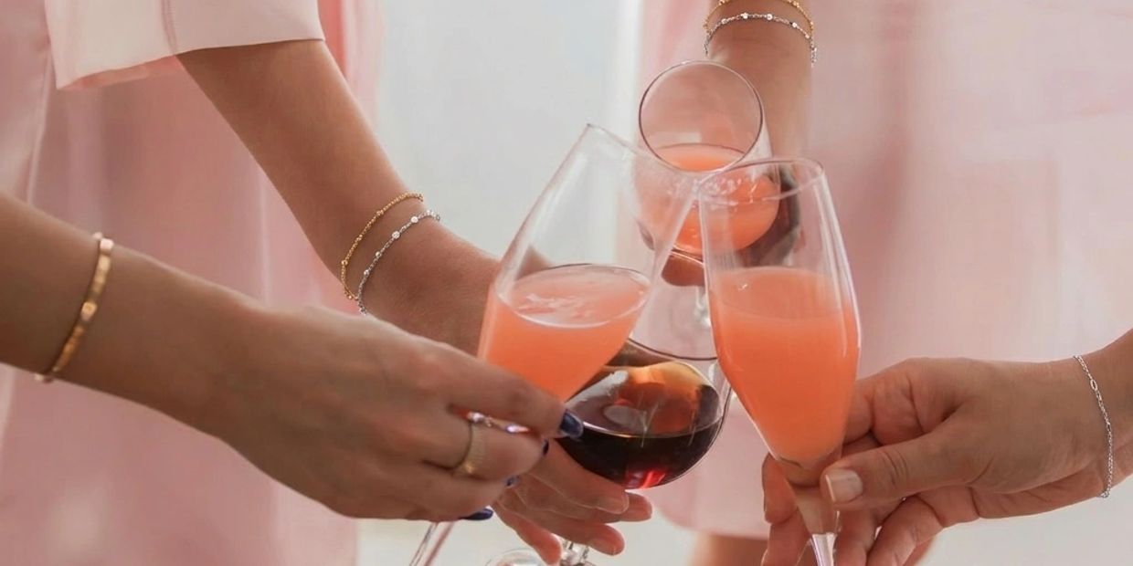 Women wearing gold and silver permanent jewelry bracelets while clinking wine glasses.