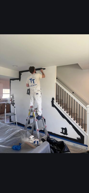 Painter applying black paint on a white wall near staircase.