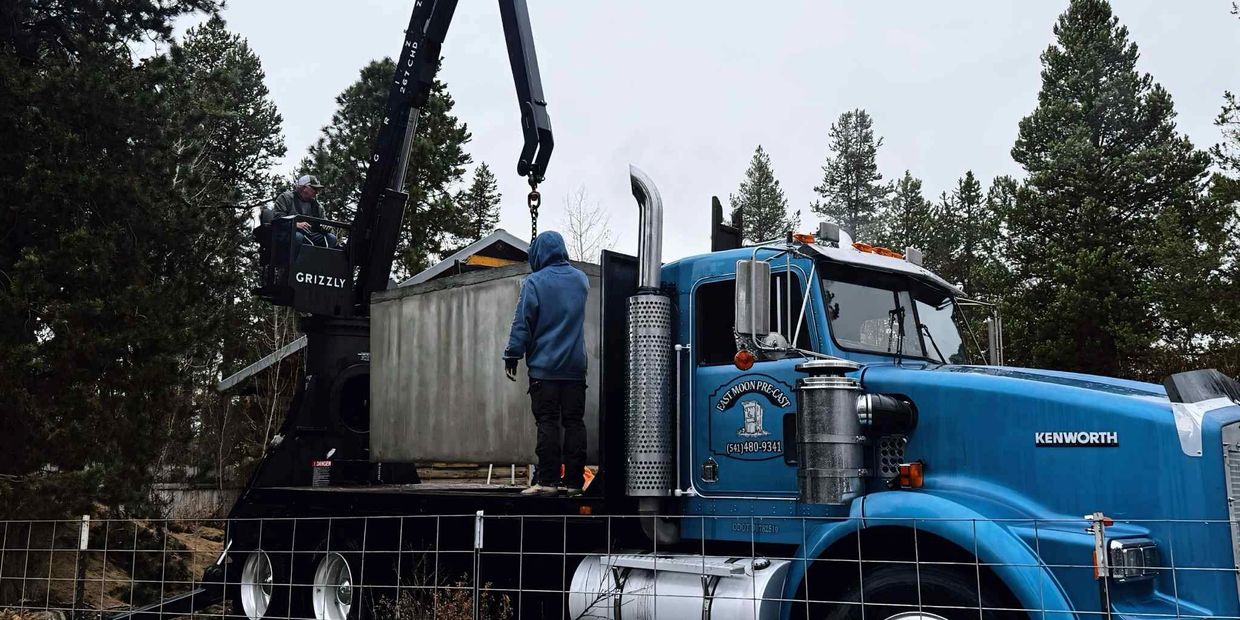 A blue Kenworth truck with a crane lifting a concrete structure in a forested area.