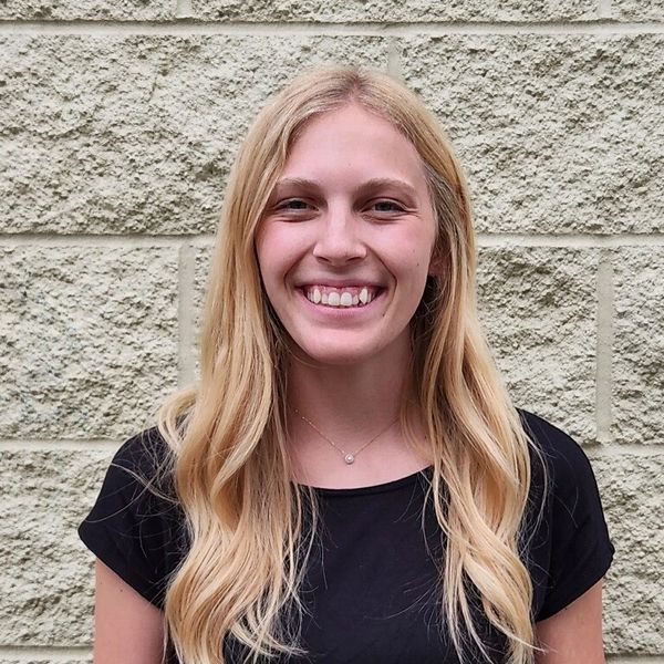 Young woman with long blonde hair smiling against a stone wall.