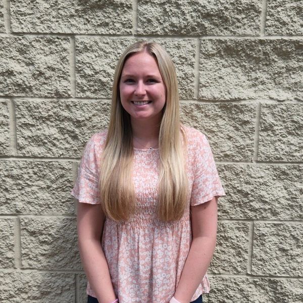 Smiling woman with long blonde hair stands against a textured brick wall.