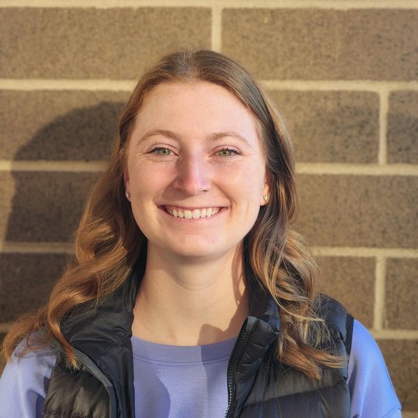Smiling woman in a black Patagonia vest and purple shirt against a brick wall.