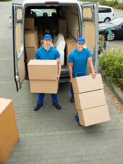 Two male movers in blue uniforms smiling while unloading cardboard boxes from the back of a van.