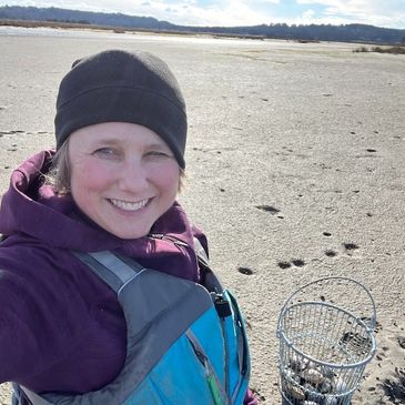 A photogaph of a smiling woman in a hat and lifejacket on a beach at low tide.