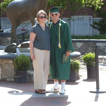 A photograph of a mother and son in front of a ram statue. The son is wearing a gradate cap and gown