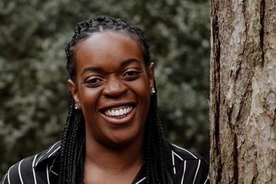 Divina Johnson in a black and white striped shirt leaning against a tree trunk