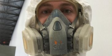 Person wearing white protective suit and 3M respirator mask in an industrial setting.