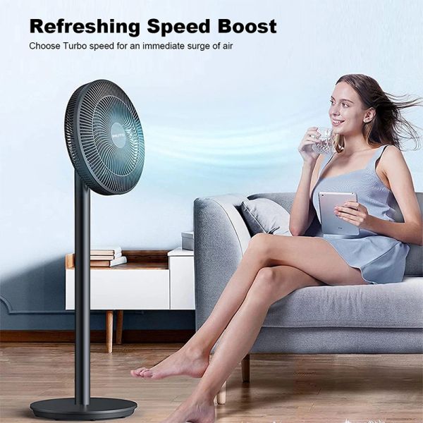 Woman enjoys cool breeze from a modern standing fan while relaxing with a tablet.
