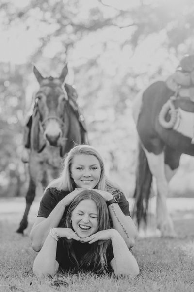 Two women posing happily on grass with horses in the background.