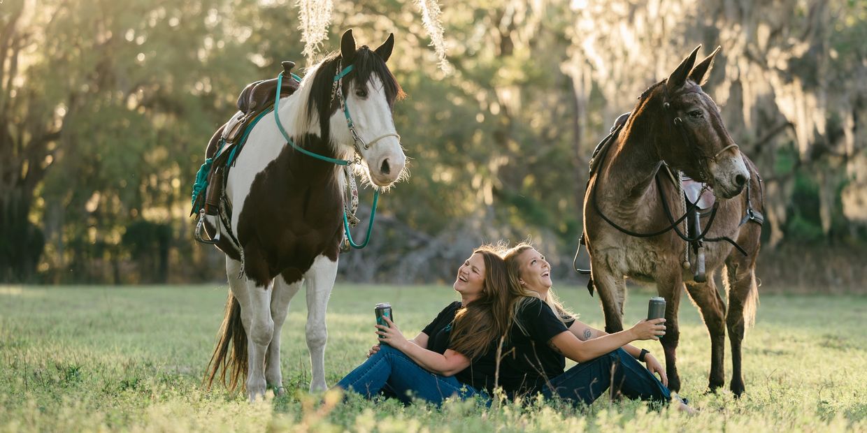 Two women sit back-to-back in a field with two saddled horses beside them.