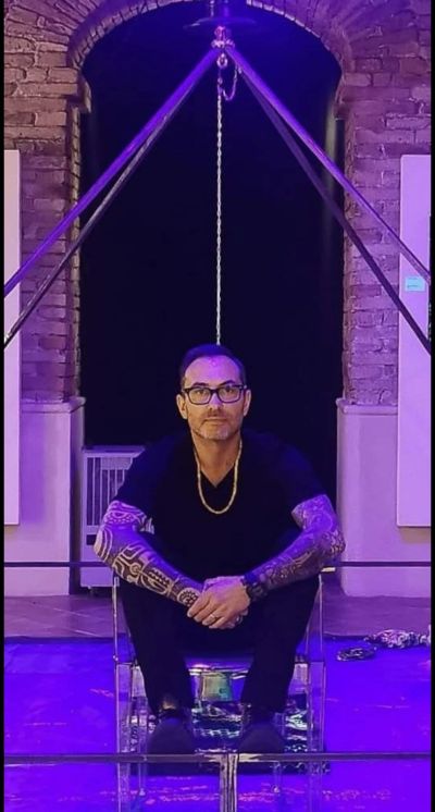 Man with arm tattoos sitting under a large tripod structure.