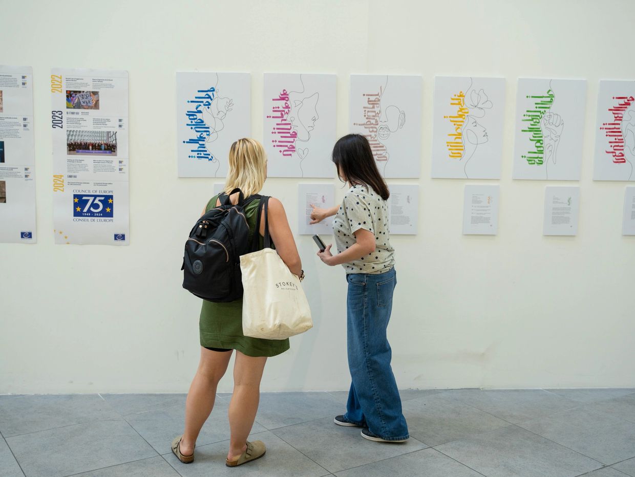 Two women observe and discuss artwork displayed on a white gallery wall.