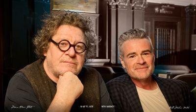 Two men sitting indoors, one with curly hair and round glasses.