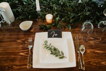 A wedding place setting with a white plate, silver cutlery, a wooden harvest table, and greenery.