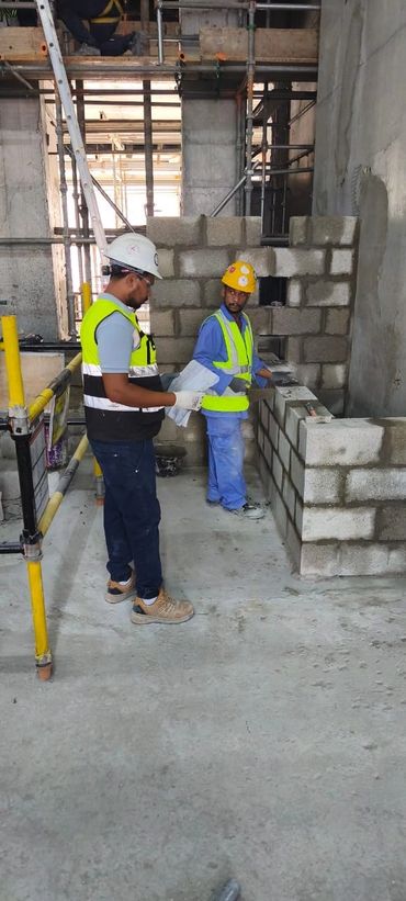 Two construction workers in safety gear working on a concrete block wall inside a building under construction.