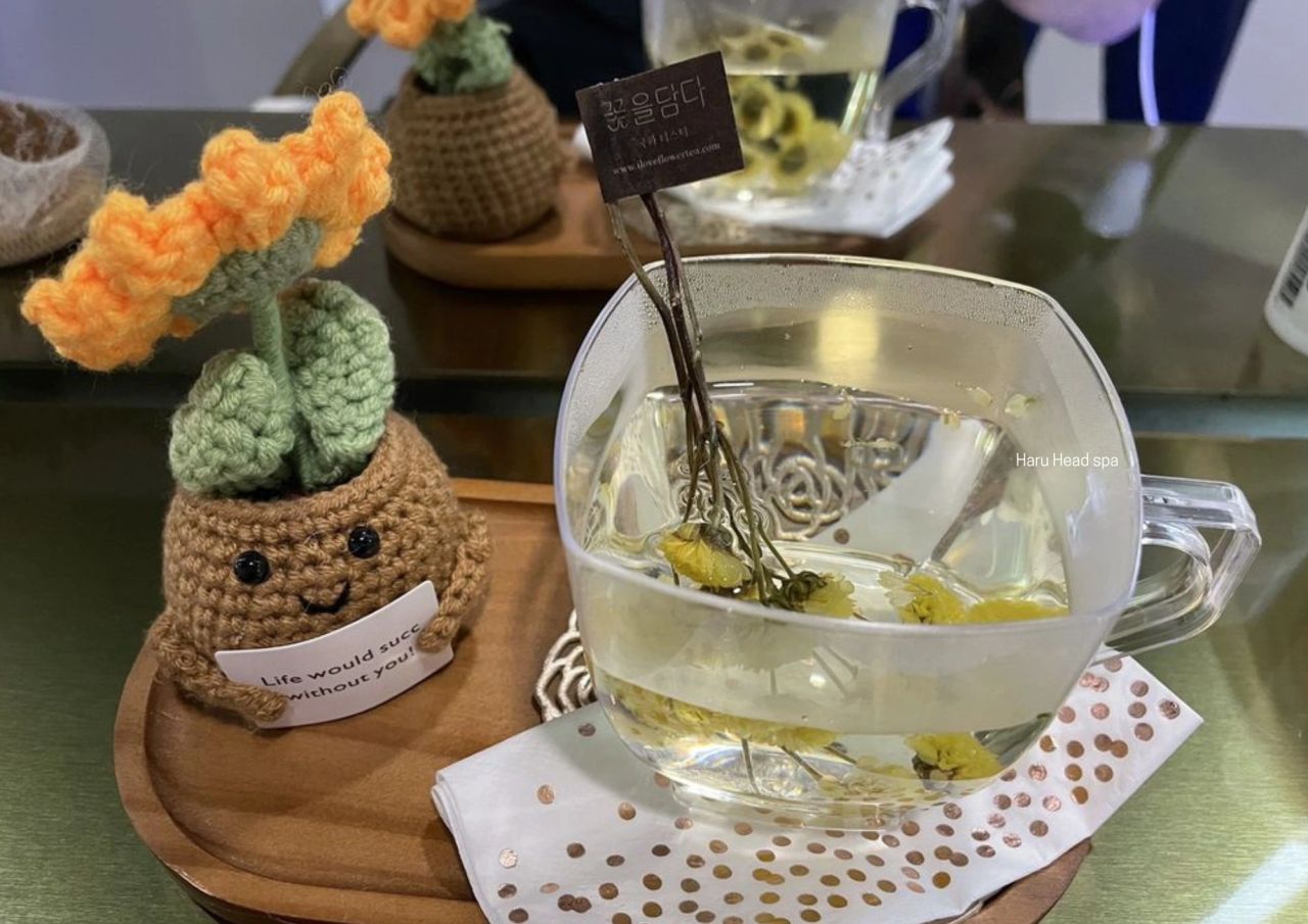 A clear glass cup of herbal tea with floating flowers, placed on a wooden tray with a crocheted flower pot and a sign that reads 'Life would suck without you!'