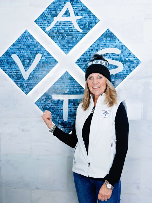 Woman in white vest and black beanie stands by a blue mosaic wall with letters.